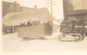 On 12th ave just north of 4th st, Princess Cafe on right, small building behind the Empire Hotel the rear of which is shown on the left. International Harvester Crawler Tractor pulling a large wagon style sleigh, full of local folks 